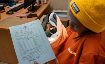 A young woman in a yellow cap and orange shirt sits by a desk holding her baby in her lap and a piece of paper in her hand toward the camera.