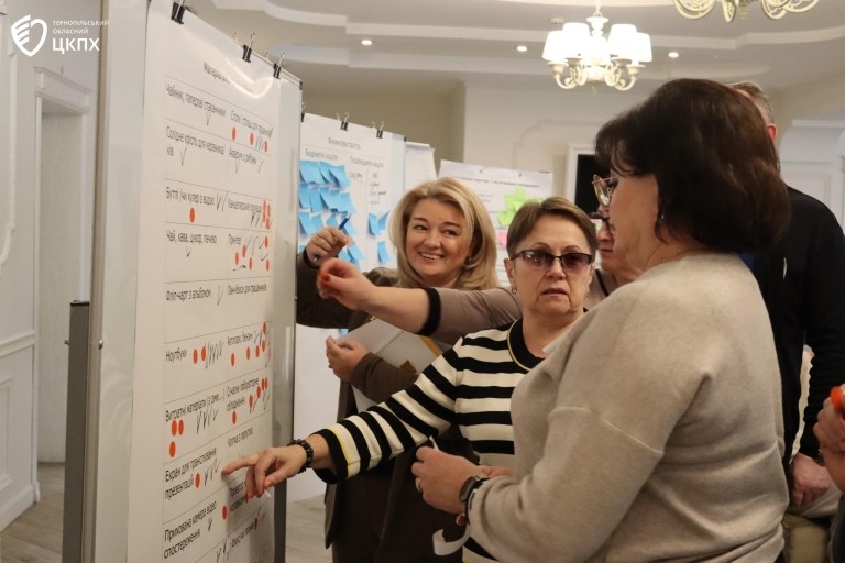 Three women stand inside looking at a poster and having a discussion about its content.