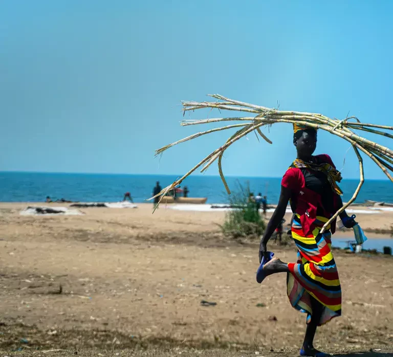 A woman walks along a beach by Lake Malawi with clear blue skies overhead. Credit: Brian Clark/Pact.